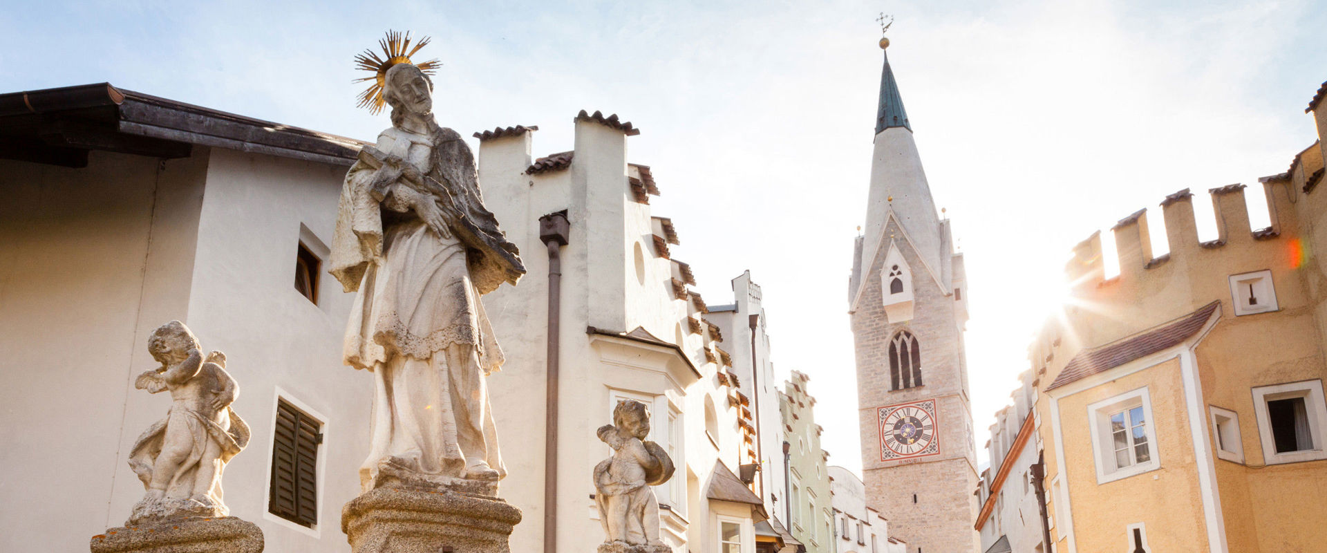 Old town of Brixen. Statue of a saint flanked by two angels on a stone arch, behind it pastel-coloured house facades and the church tower of an old town.