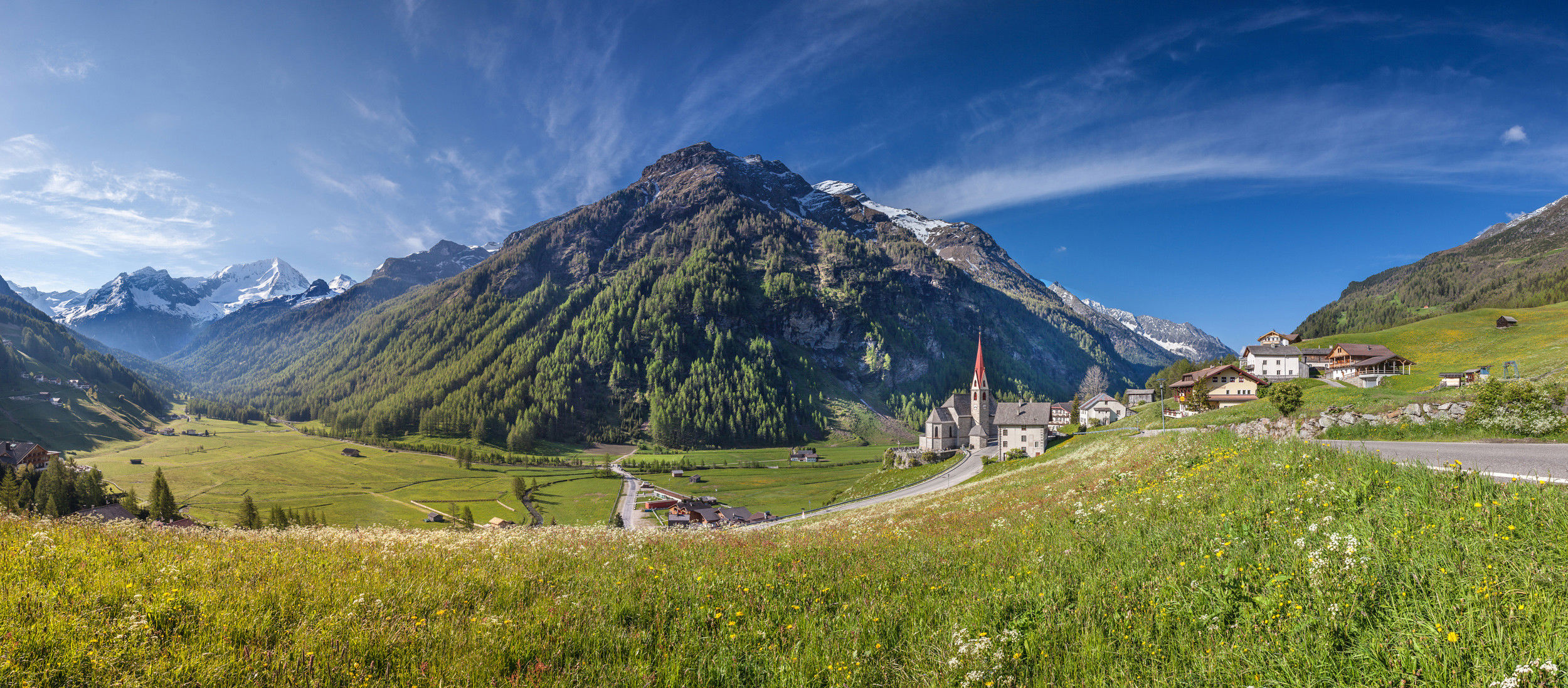 View of valley floor, church and houses, framed by flowering mountain meadows.