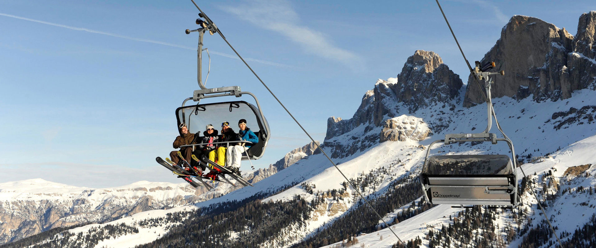 Winter in Eggental. Four skiers in a chairlift in front of a mountain landscape with blue sky.