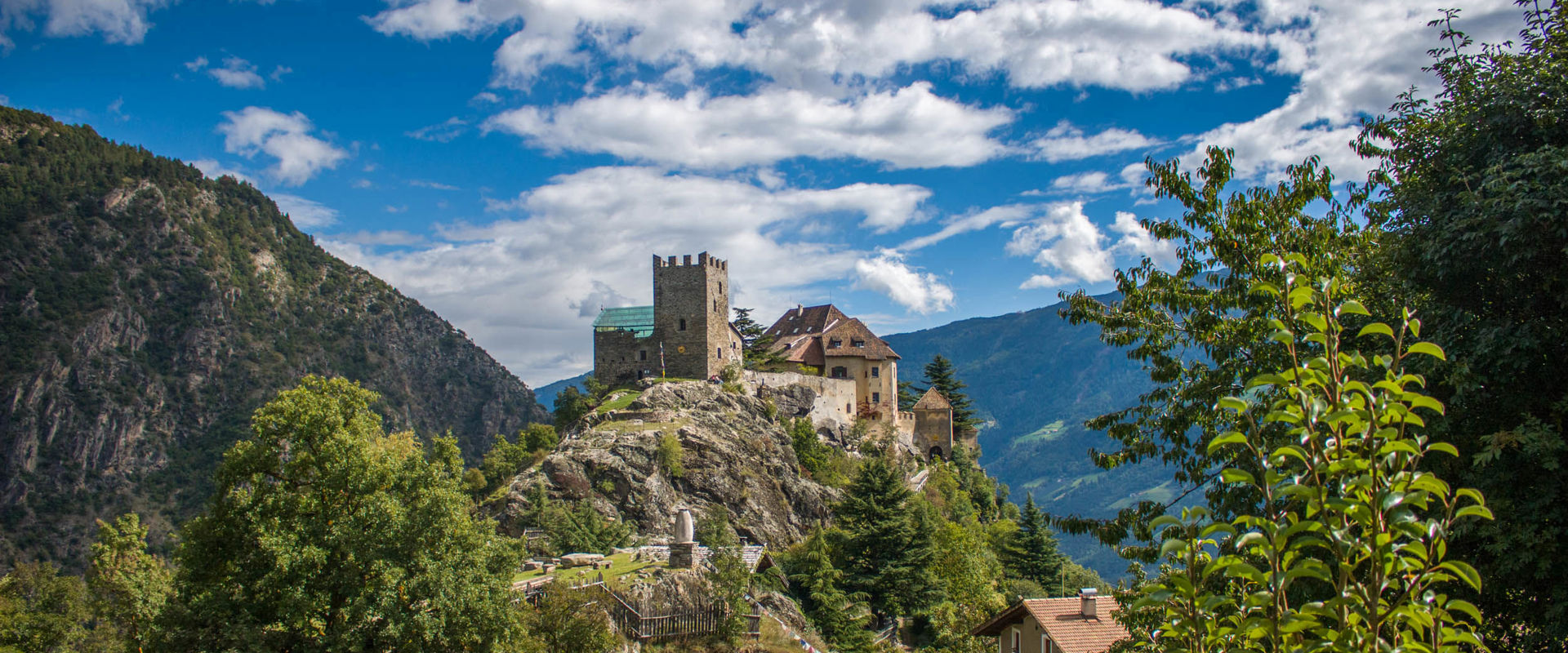 Juval Castle The Juval Castle is perched on a rocky hill surrounded by trees, mountains, a house and a small hut.