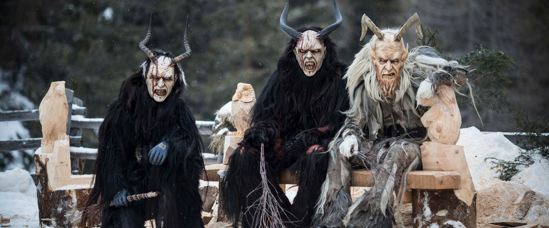3 people dressed up as Krampus sit on a wooden bench and look directly into the camera.