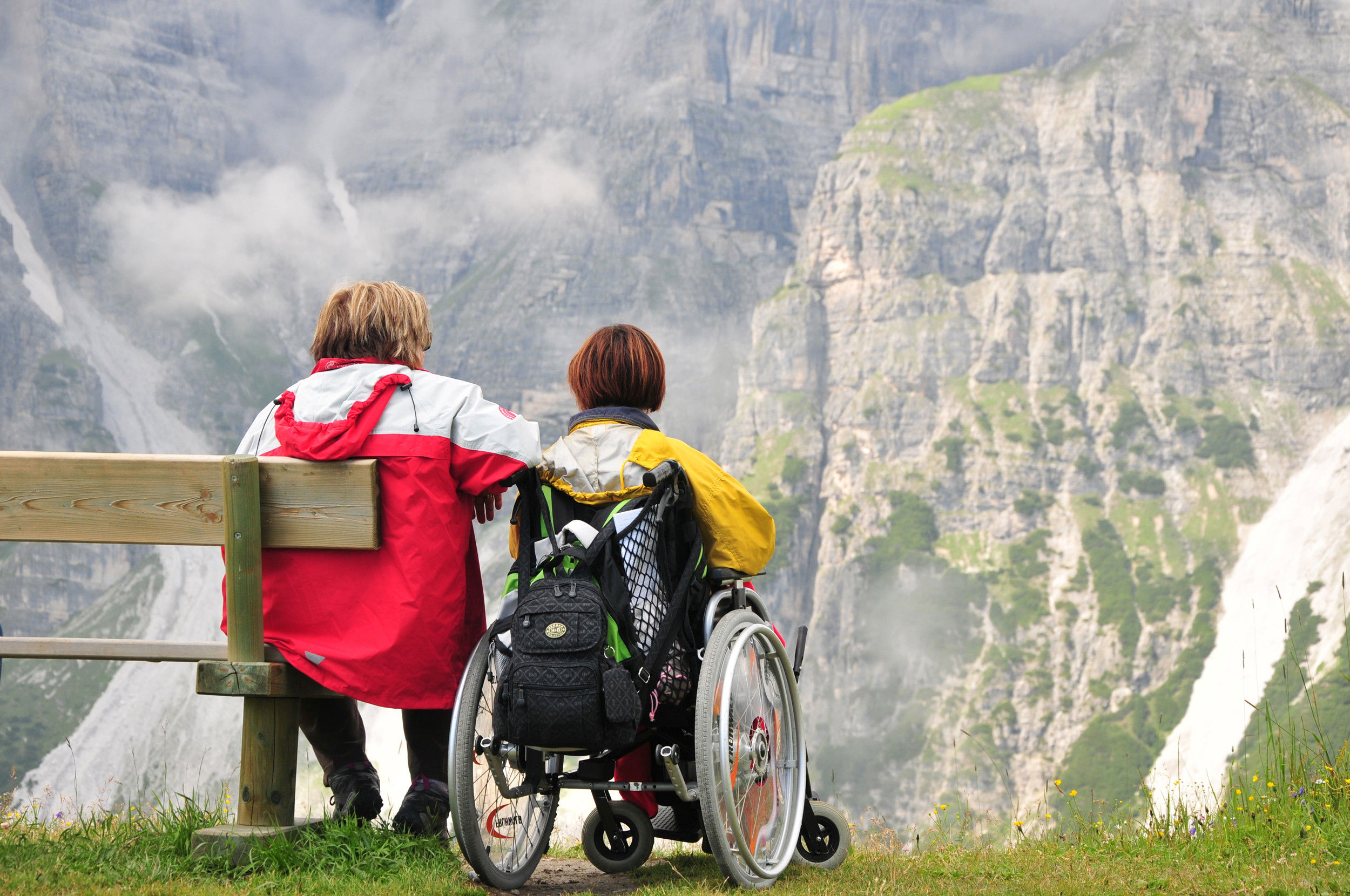 A man is sitting on a wooden bench, while the woman next to him is sitting in a wheelchair.
