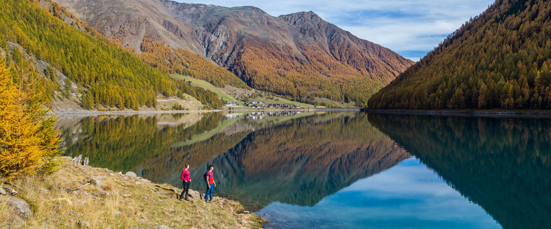Young couple standing on the shore of the Vernagt reservoir. The surrounding mountains are reflected in the water.