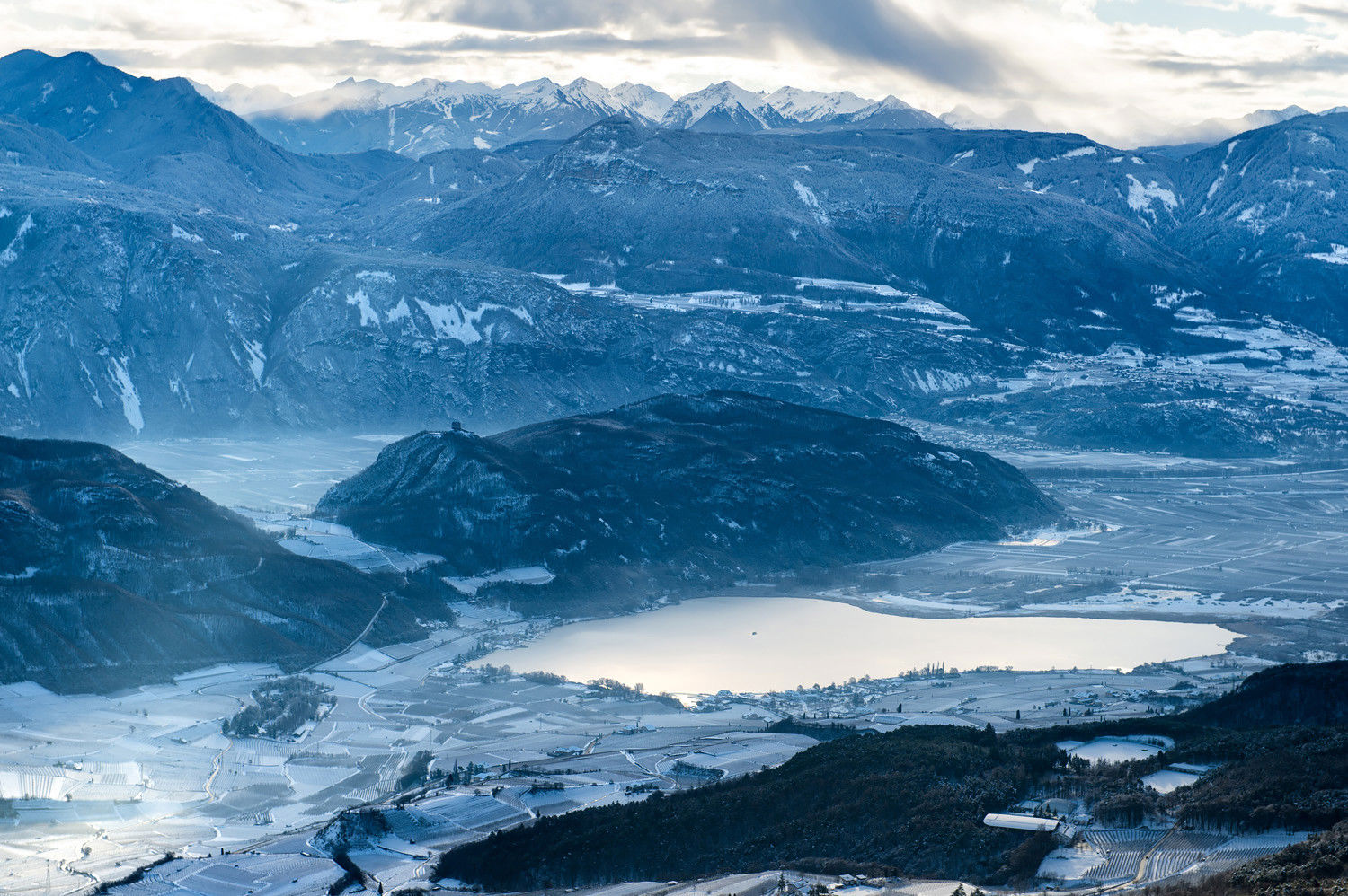 Panoramic view of Lake Kaltern in winter. The vineyards, mountains and surrounding nature are covered in snow. 