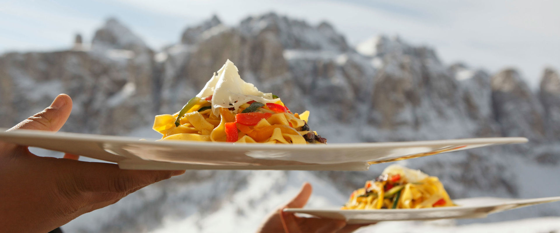 Eating with a wintery atmosphere. A waiter serves 2 pasta dishes in front of a wintry mountain backdrop.
