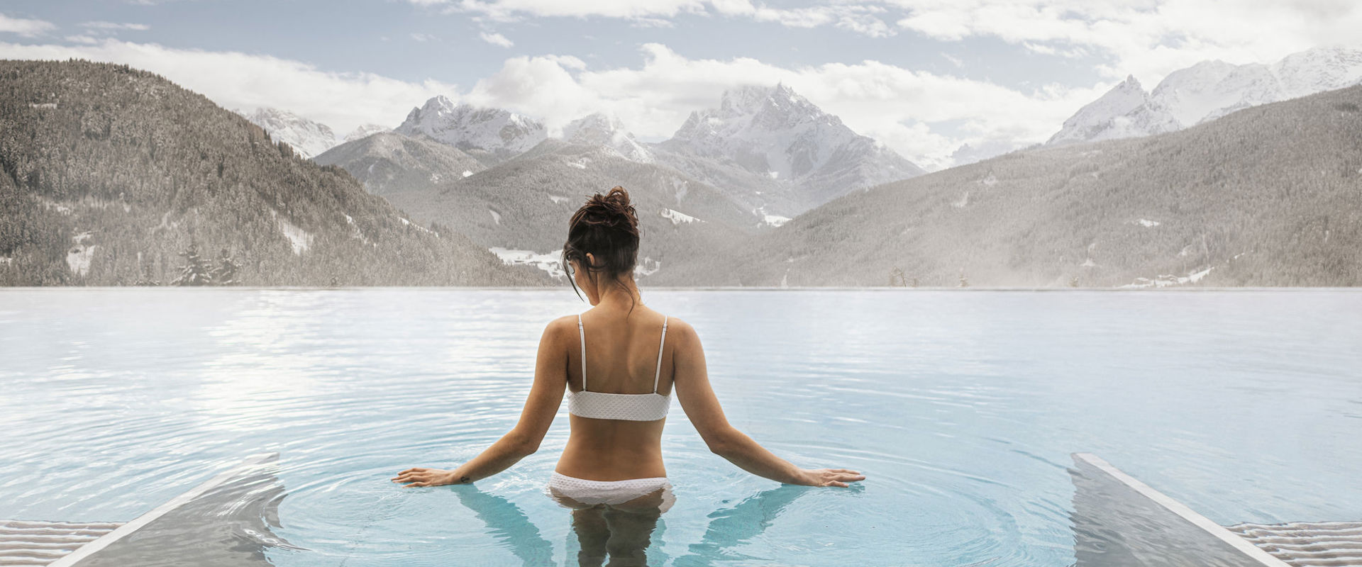 Woman wearing a white bikini and with dark hair tied up, descends the steps into the steaming outdoor pool. In the background, you can see the snow-covered mountains.